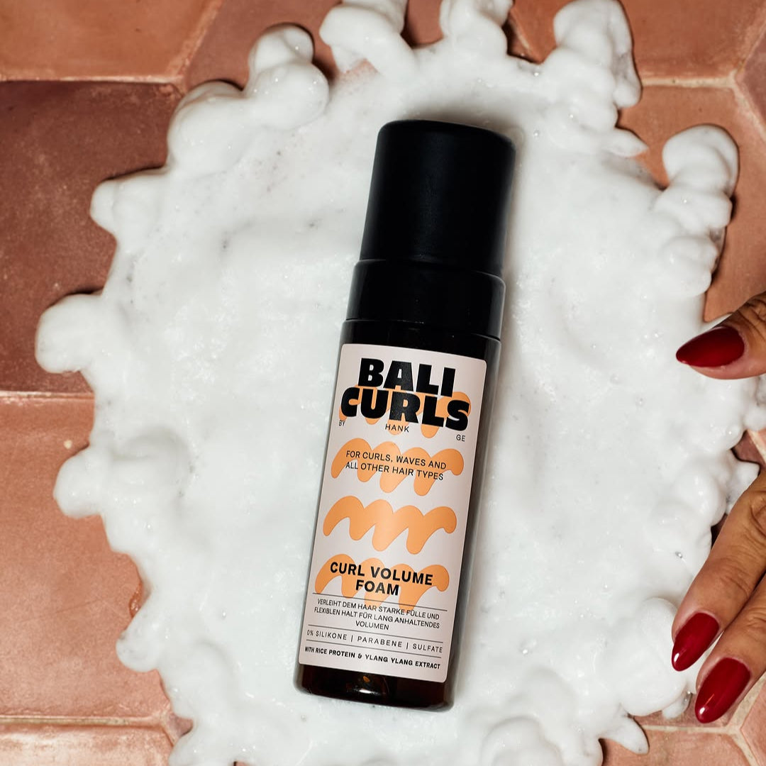 Bottle of Bali Curls foam on a pile of white foam with a hand holding it, on a tiled floor.