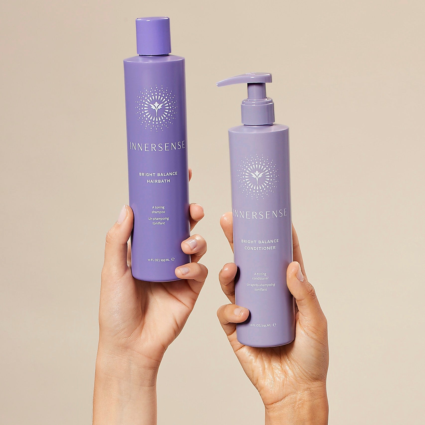 Two hands holding purple bottles of Innersense hair care products against a beige background
