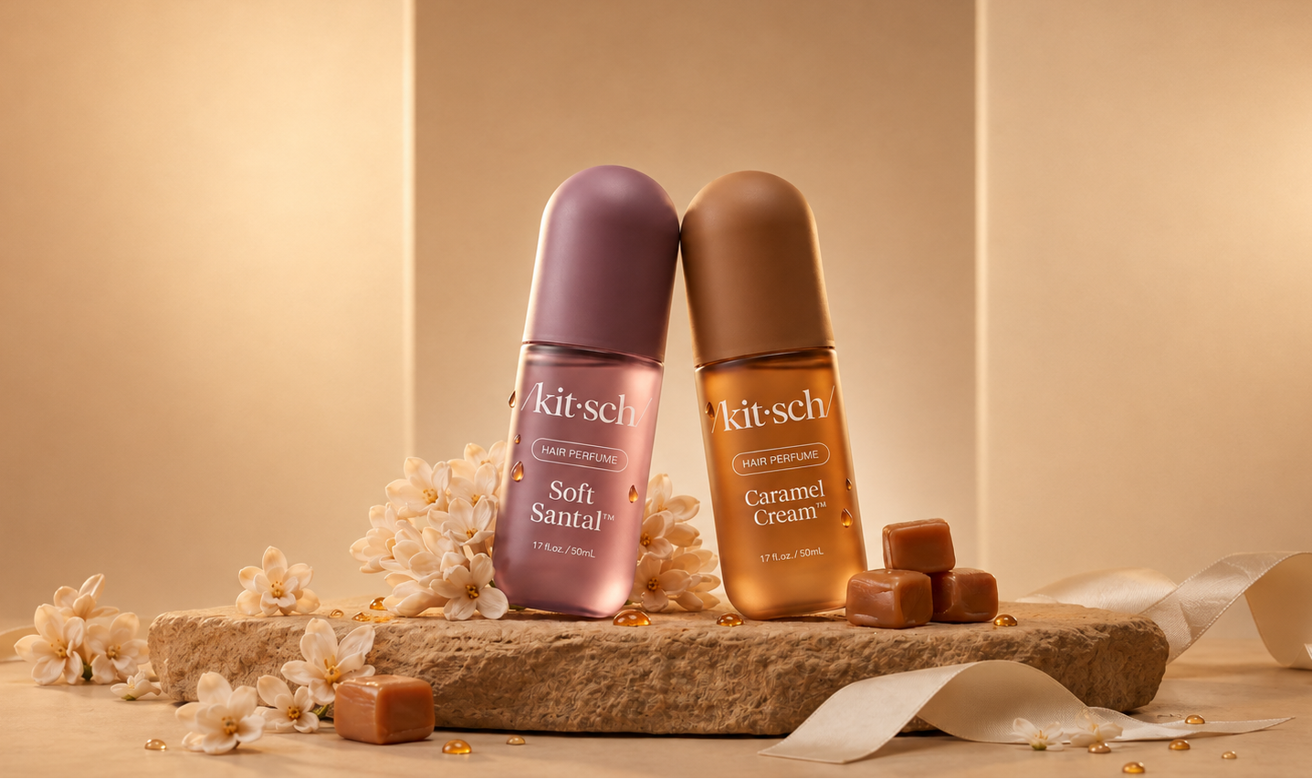 Two Kitsch deodorant bottles, one labeled Soft Sandal in a dusty purple shade and the other labeled Caramel Cream in a warm brown shade, displayed on a stone slab with caramel pieces, dried flowers, and white ribbon against a warm beige background.