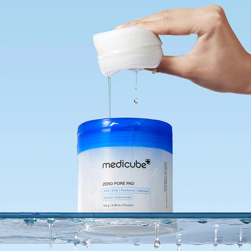 Hand squeezing a white pad over a jar of Medicube Zero Pore Pad on a blue background