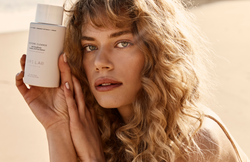 blonde curly hair on a beige background holding a bottle of ori lab cleanse - kiyo beauty