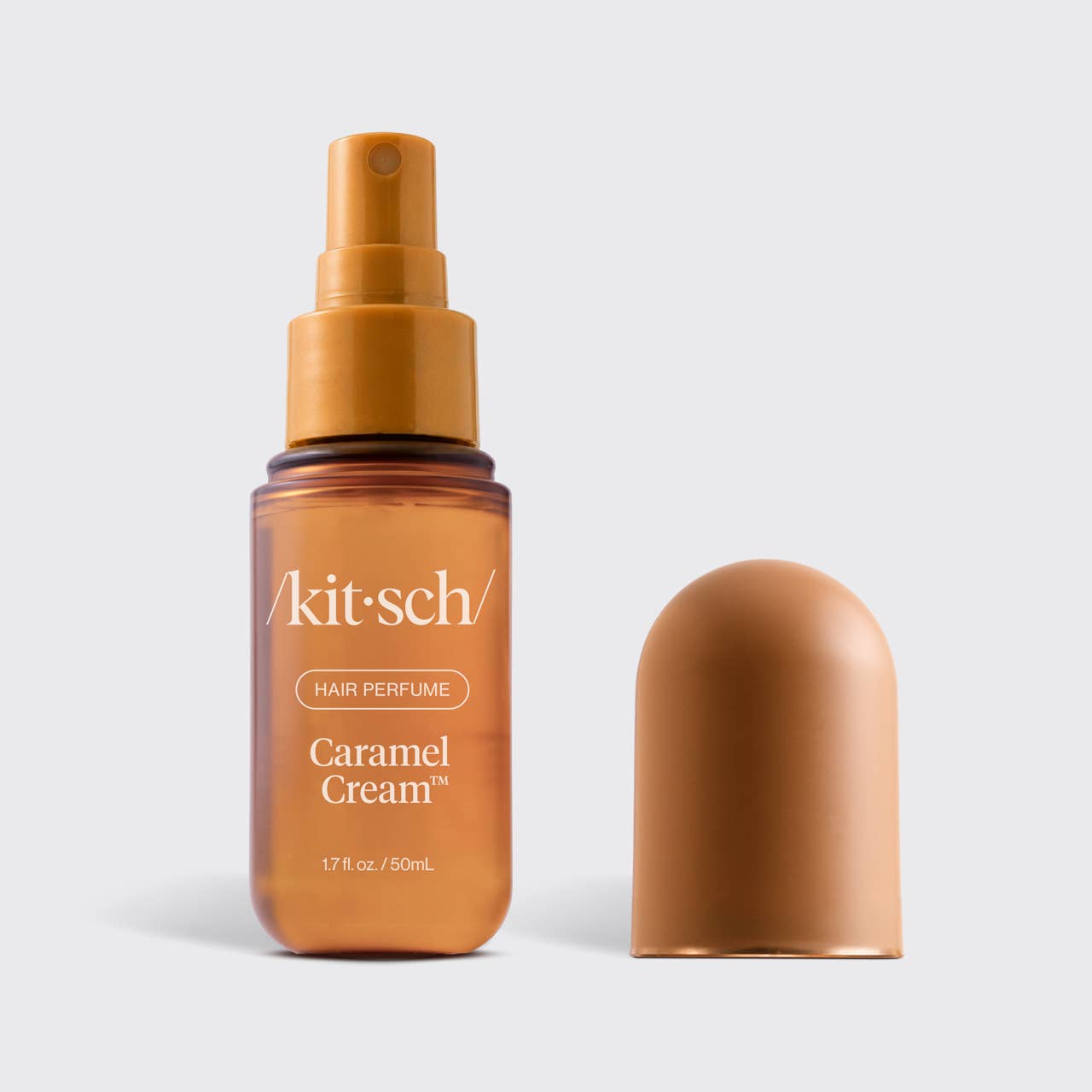 A bottle of Kit-Sch Hair Perfume labeled 'Caramel Cream' in an amber-brown glass bottle with a matching brown spray top, shown next to its detached cap on a light gray background.