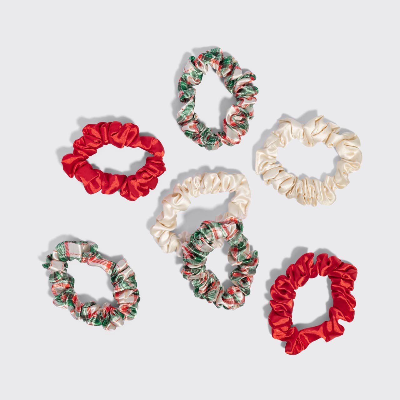 Six satin hair scrunchies arranged on a light gray background, featuring solid red, cream, and green-and-red holiday patterned designs.