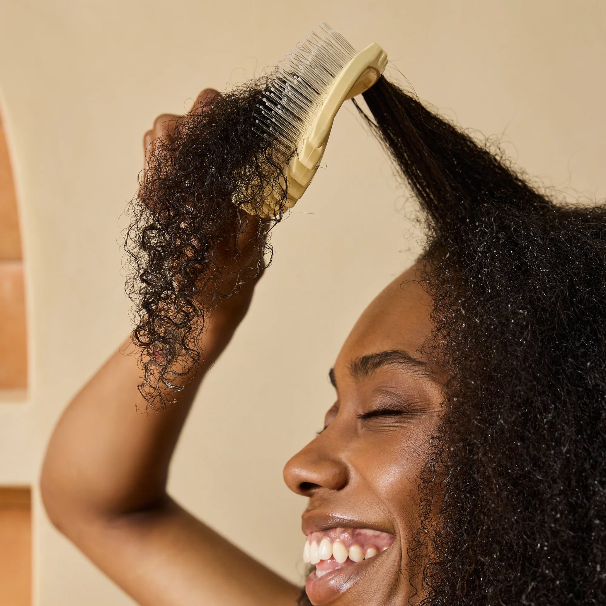 A smiling woman with dark curly hair uses a cream-colored detangle edgelift bounce curl detangling brush to brush her hair, shown in a close-up profile view against a warm beige background.