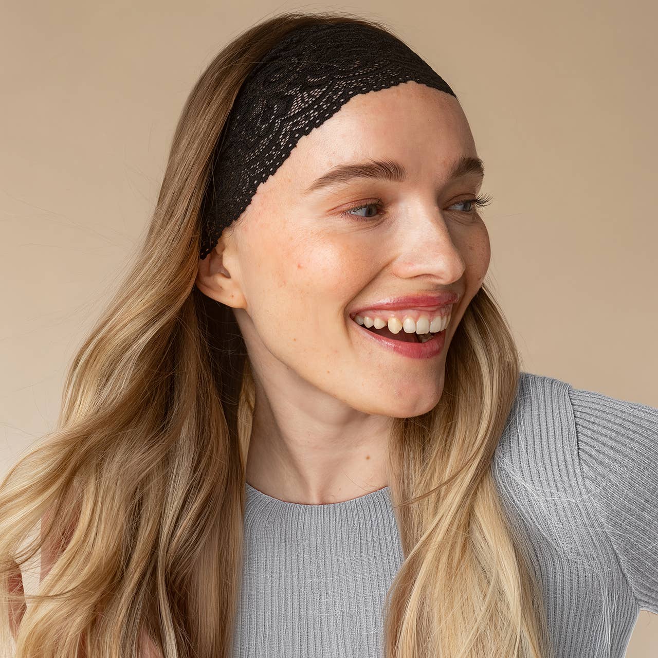 A woman with long blonde hair wearing a kitsch black lace headband and a gray ribbed top, smiling against a solid beige background.