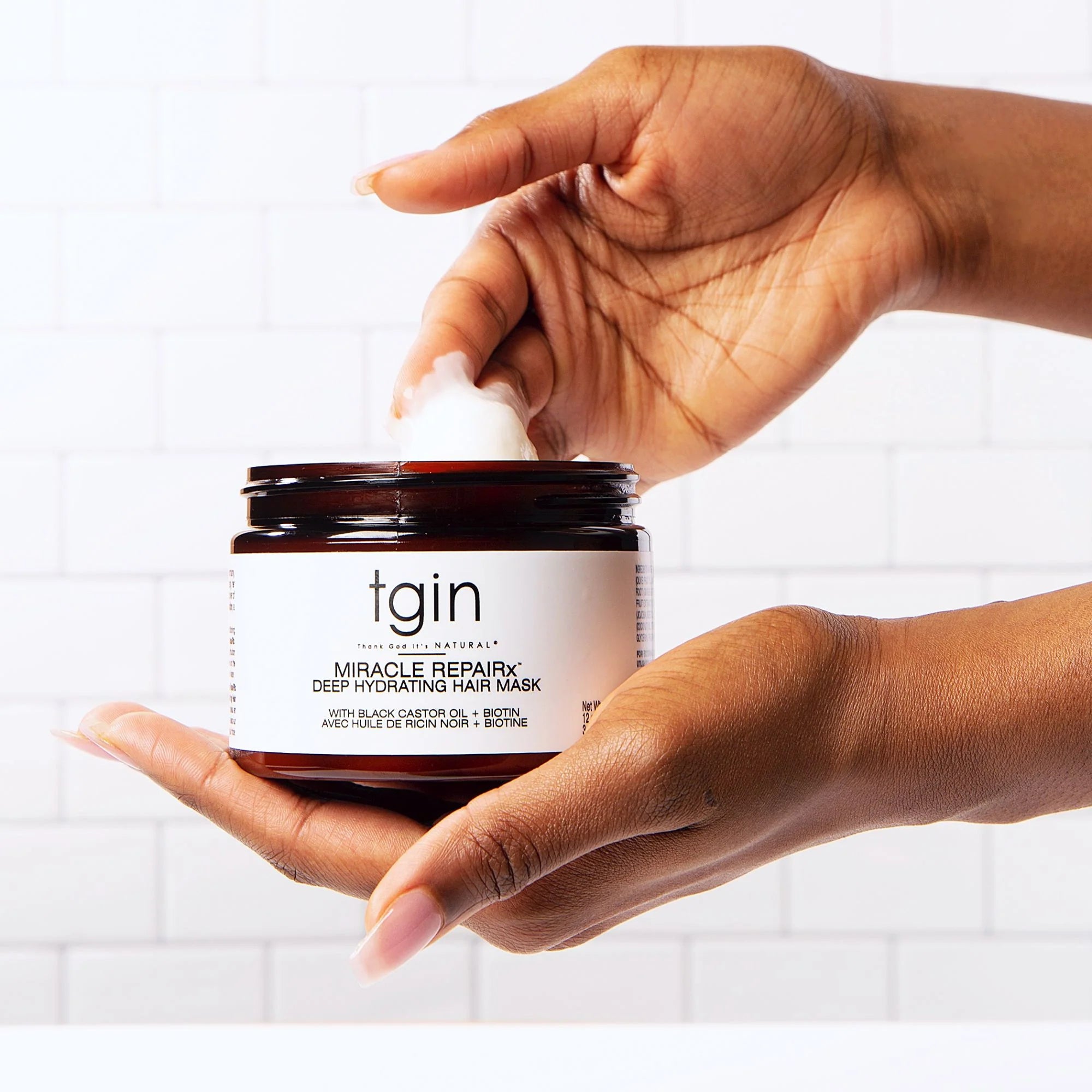 A person's hands holding and scooping from a jar of tgin Naturals Miracle Repair Deep Hydrating Hair Mask against a white subway tile background - Kiyo Beauty