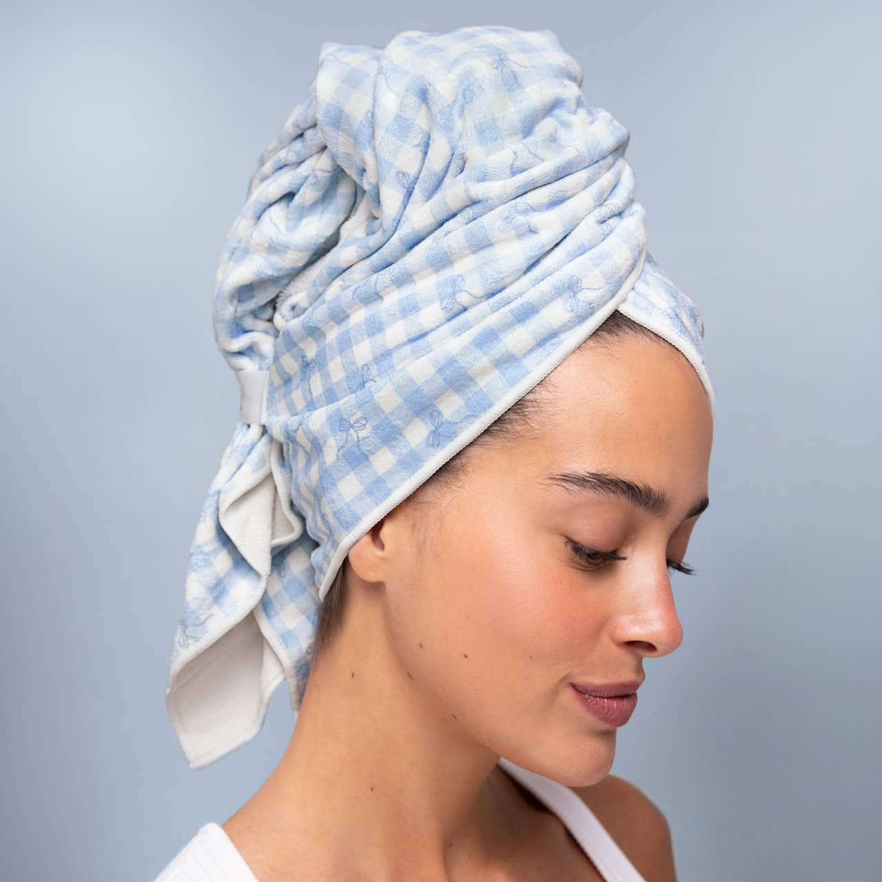 A woman wearing a light blue and white gingham checkered cotton towel wrapped around her head, shown in profile against a soft light blue-gray background. Microfibre Hair Towel | Blue Gingham Hair Wrap | Kiyo Beauty