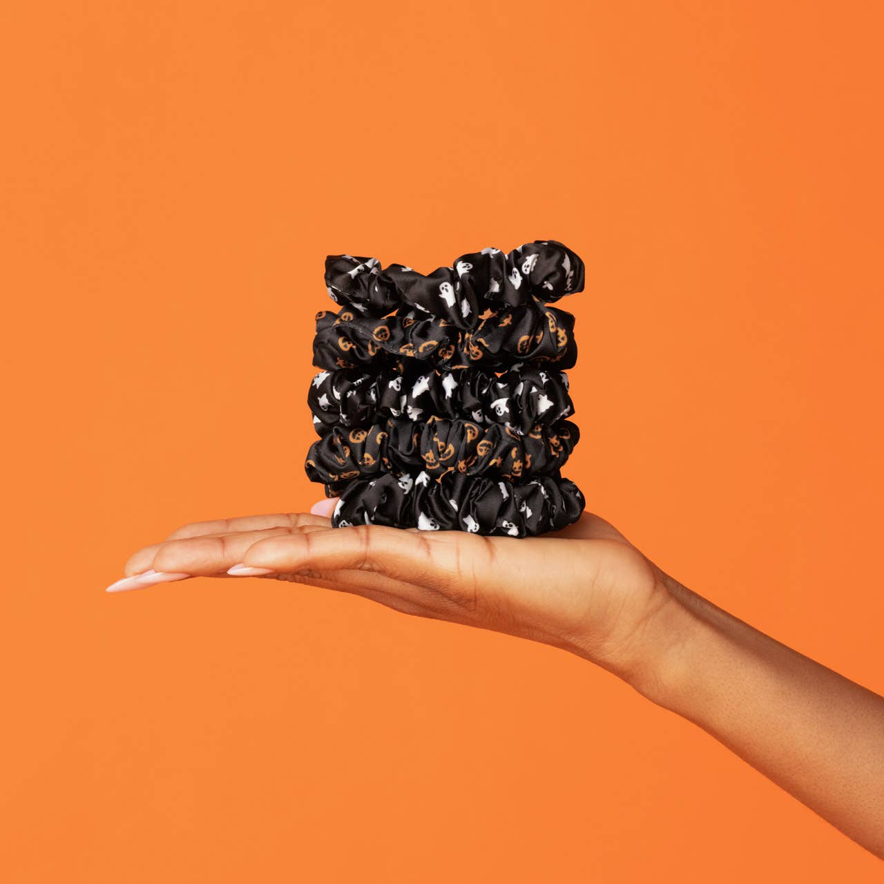 A hand holding a stack of black satin scrunchies with a small orange and white pattern, set against a solid orange background. Kitsch Ghost & Pumpkins Scrunchies