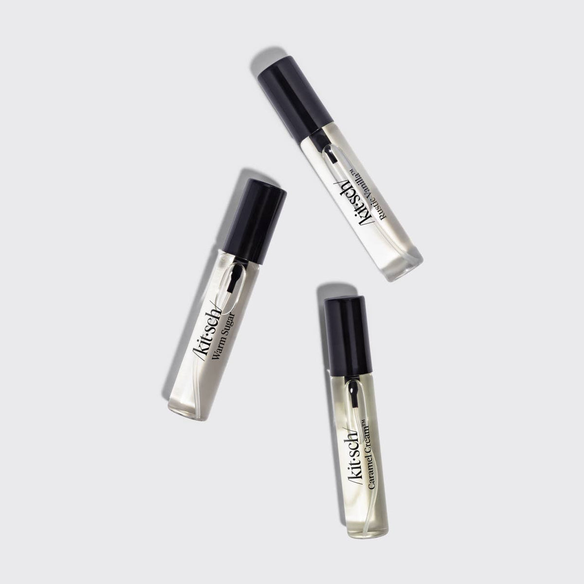 Three small clear cosmetic tubes with black caps arranged diagonally on a light gray background. The tubes are labeled 'Kitsch' and appear to be perfume mists