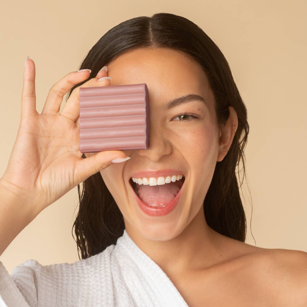 Woman holding a bar of soap in front of her eye on a beige background.Kitsch Amber Shores Body Wash Bar 

