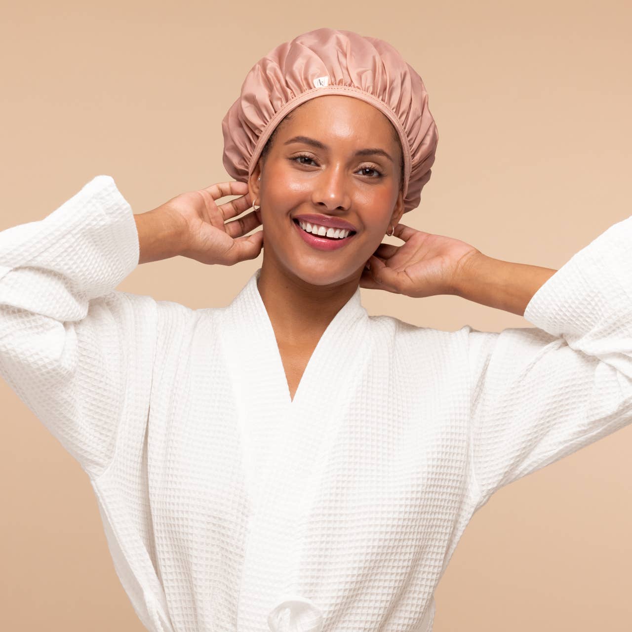 A smiling woman wearing a white waffle-knit robe and a dusty pink satin shower cap, posing with her hands behind her head against a solid beige background.