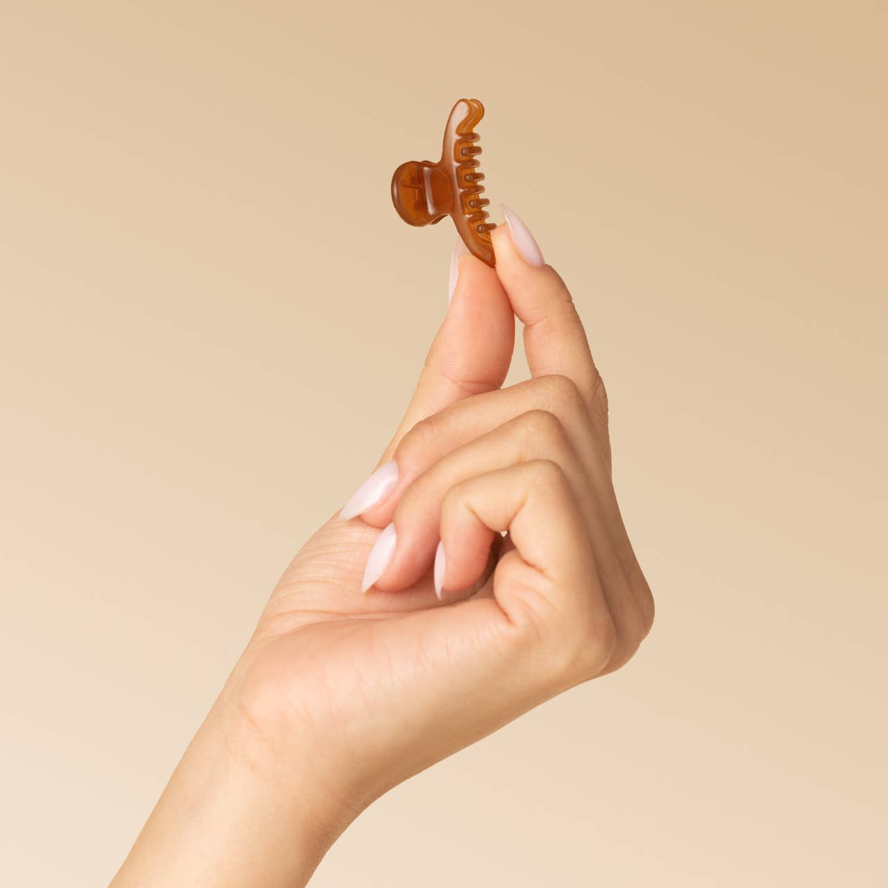 A hand with pale pink manicured nails holds a small amber-brown hair claw clip against a solid warm beige background.