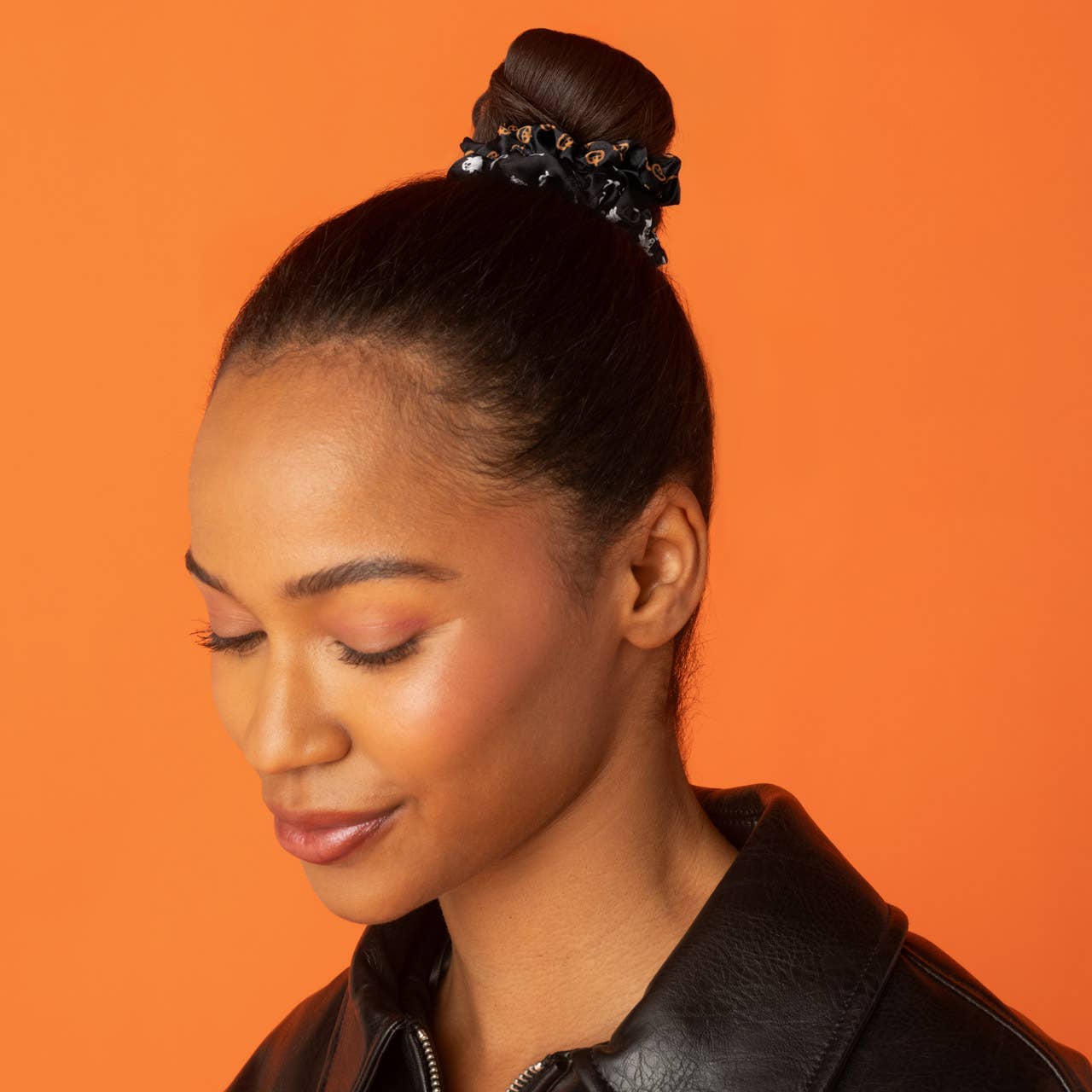 A woman with her hair styled in a high bun wearing a black beaded hair accessory, set against a solid orange background. Kitsch Ghost & Pumpkins Scrunchies