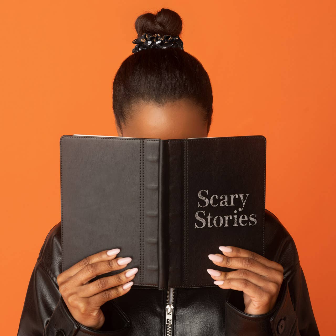 A person with dark hair in a bun holds a black book or binder labeled 'Scary Stories' in front of their face against a solid orange background. Kitsch Ghost & Pumpkins Scrunchies