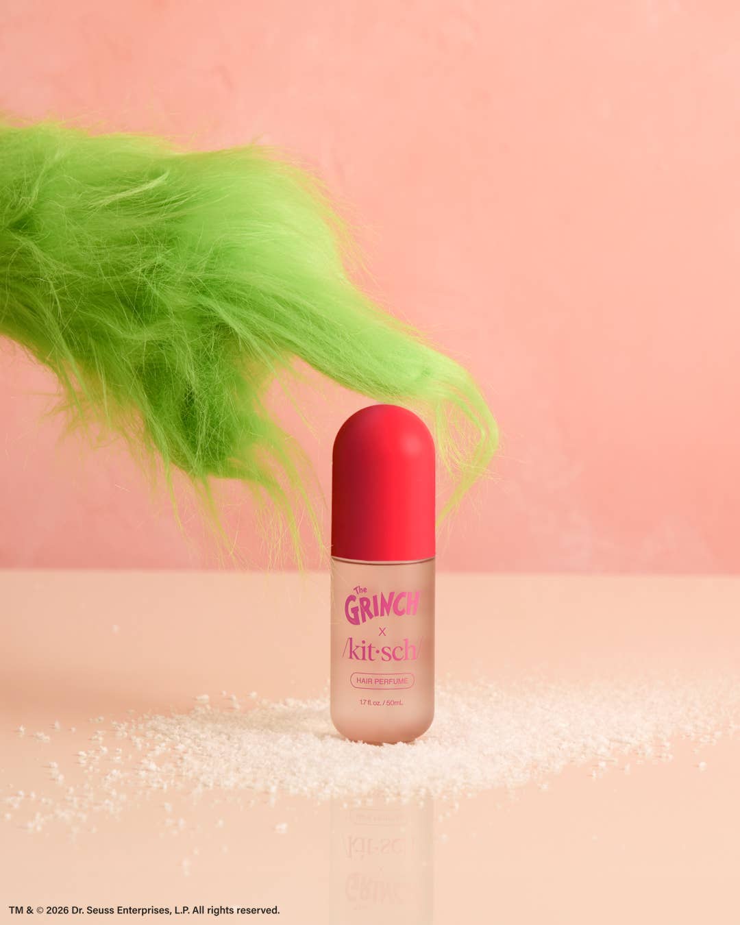 A bottle of Dr. G's Grinch & Kitsch cherry cheer hair perfume stands on a pink surface scattered with white glitter, with a bright neon green fluffy tail-like shape against a soft pink background.