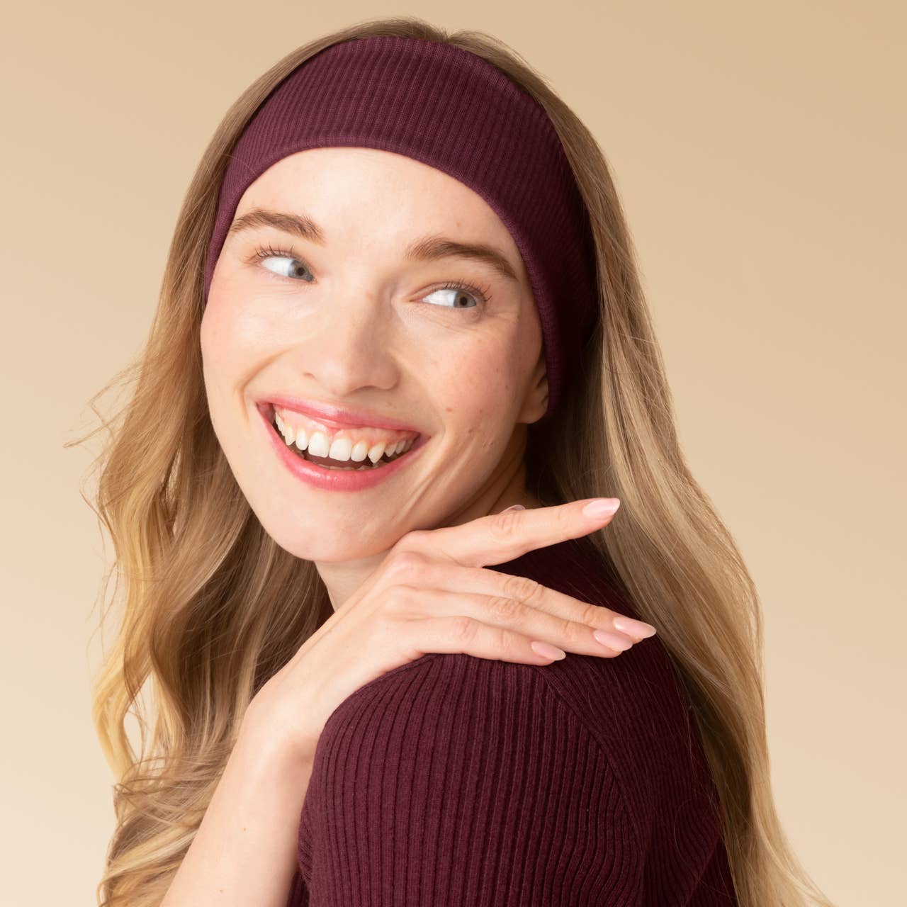 A smiling woman with long blonde hair wears a ribbed burgundy headband and matching ribbed top against a plain beige studio background.