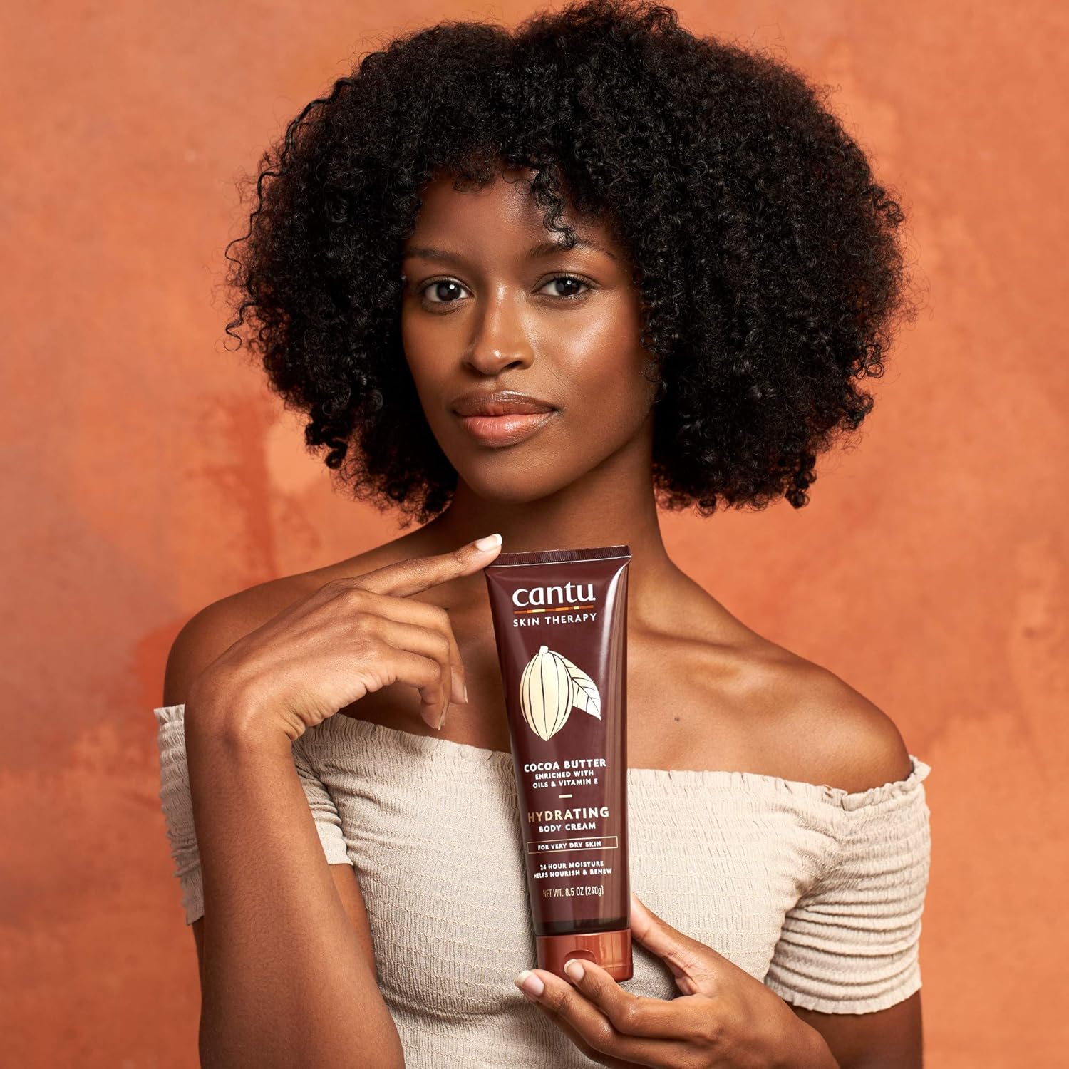Cantu Skin Therapy Shea Butter Hydrating Body Cream 240g model holding body cream tube smiling on a red sand coloured background