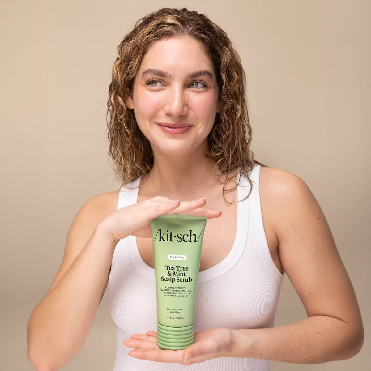 A woman with curly brown hair wearing a white tank top holds a green tube of kitsch Tea Tree Mint Scalp Scrub against a solid beige background.