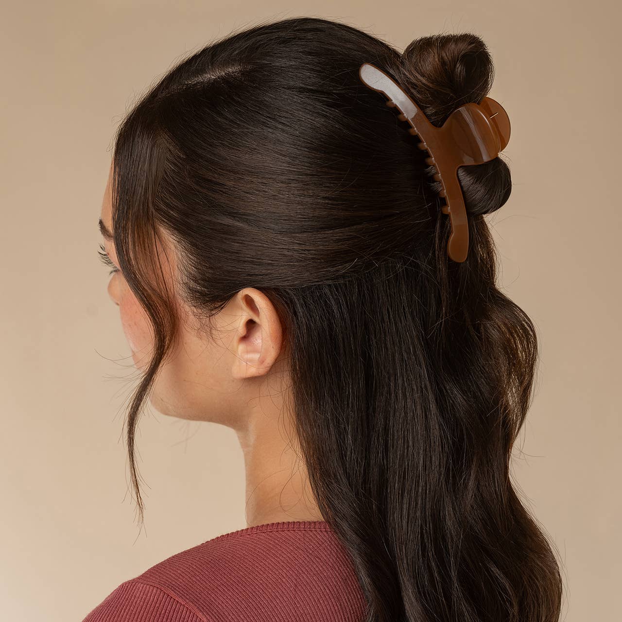 A woman shown in side profile wearing a brown translucent claw hair clip holding part of her dark hair up against a plain beige background.