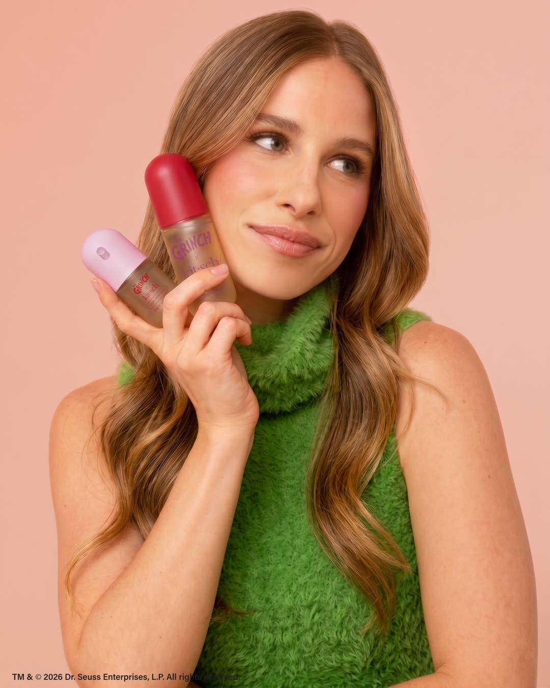A woman with long wavy light brown hair holds two Dr. Sousek cosmetic bottles against a solid pale pink background.