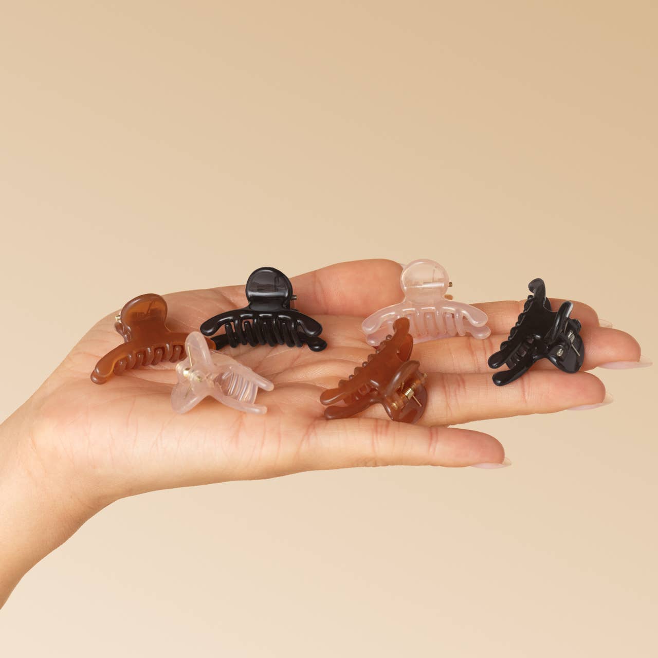 Five small hair claw clips in assorted colors including black, translucent brown, and clear, displayed on an open hand against a solid beige background.