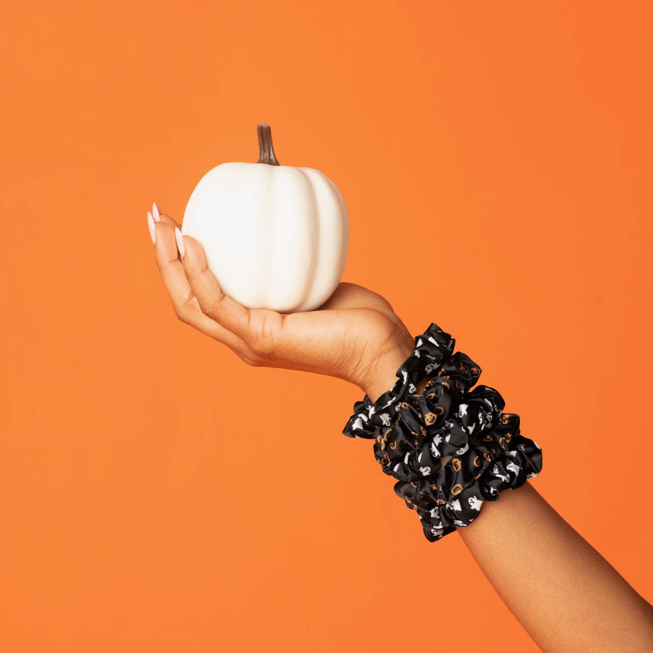 A person's hand and wrist wearing a chunky black scrunchie with white polka dots, holding a small white pumpkin against a solid orange background. Kitsch Ghost & Pumpkins Scrunchies