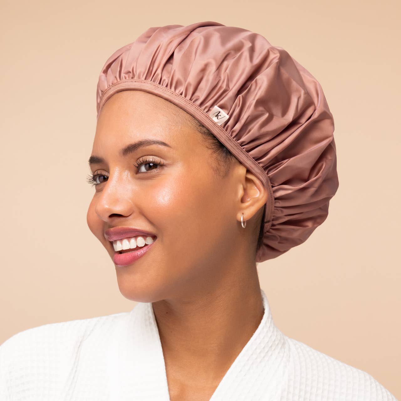A woman wearing a shiny dusty rose satin shower cap and a white textured robe, shown in profile against a solid beige background.
