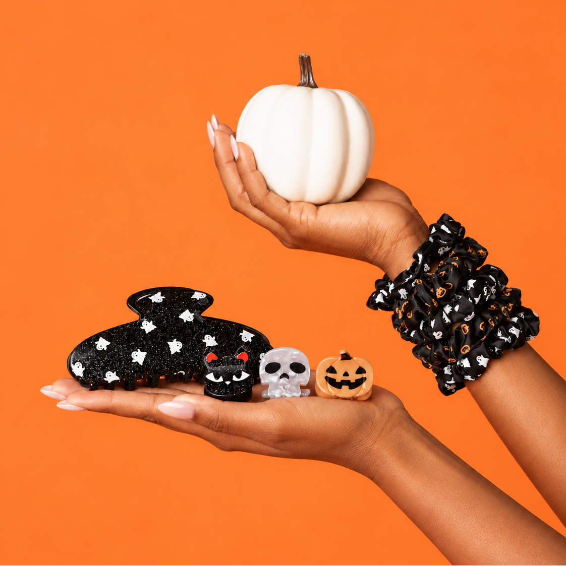 A person's hands holding a white pumpkin and small Halloween-themed accessories against a solid orange background. One hand wears a black scrunchie with white stars, and the other holds a black claw clip with white stars, a small black cat charm, a white skull charm, and a small orange pumpkin charm.