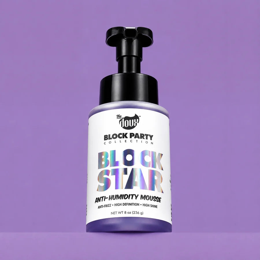 A bottle of The Doux Block Party Collection Block Star Anti-Humidity Mousse, featuring a white label with holographic lettering and a black pump dispenser, set against a solid purple background.