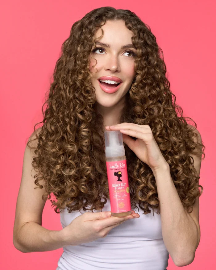 A woman with long, curly brown hair holds a bottle of Camille Rose Guava Slip Hold Up Setting Mousse 207ml | Kiyo Beauty against a solid pink background. The bottle is pink and white with a black silhouette logo.