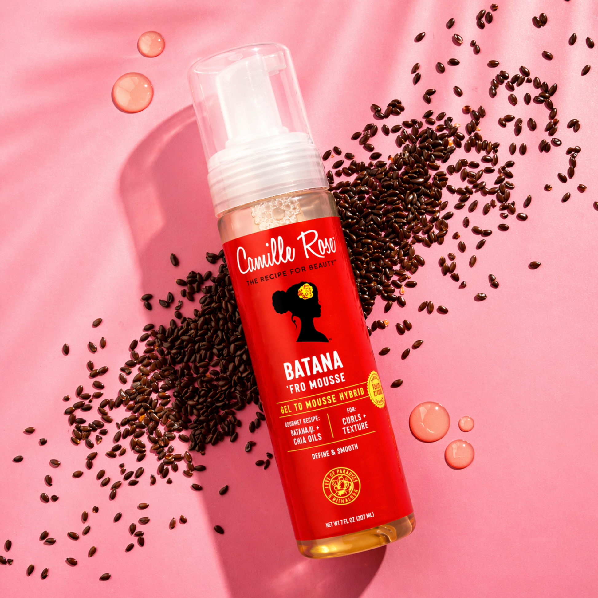 Red bottle of Camille Rose Batana Fro Mousse with black seeds on a pink background