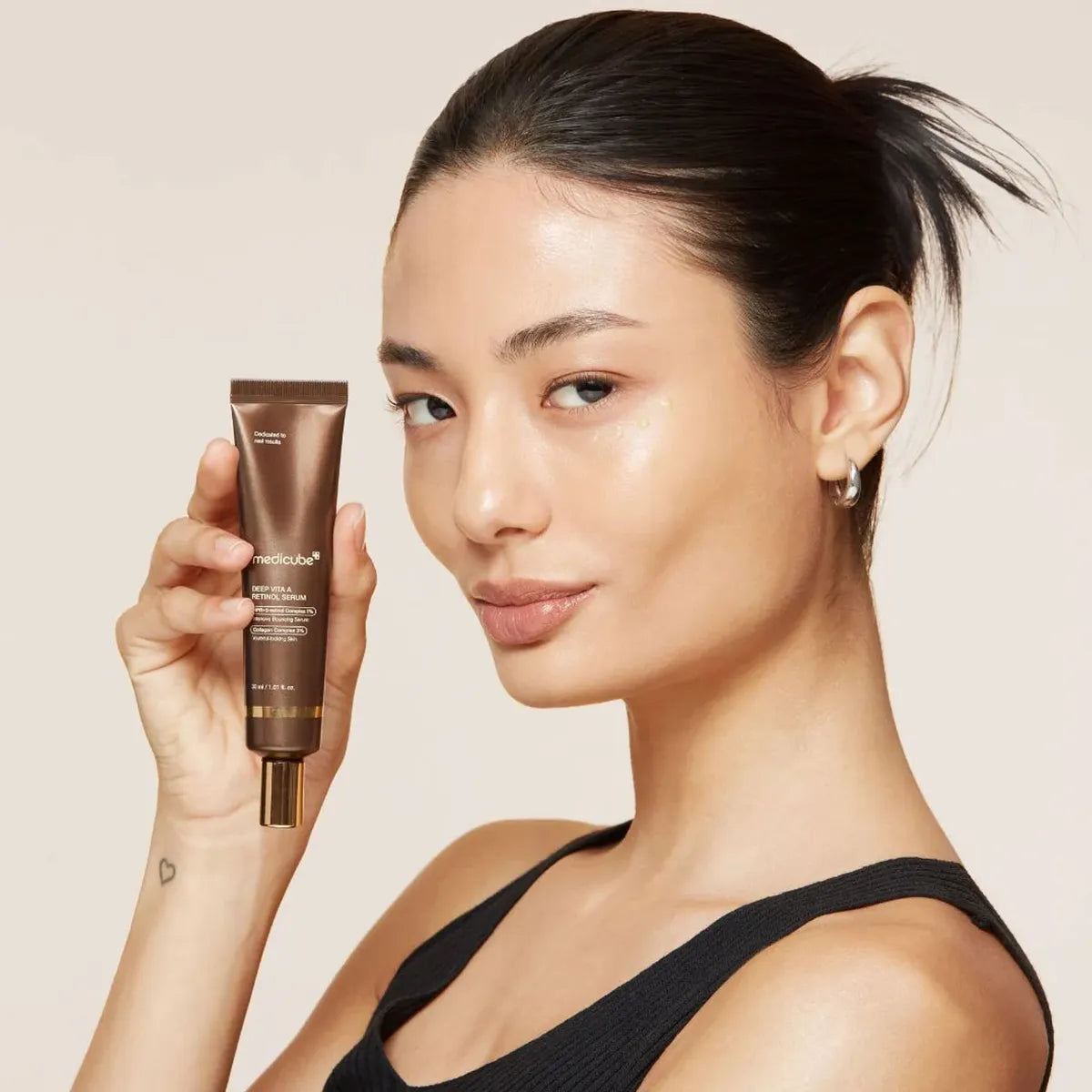 A woman with dark hair tied back holds a brown cosmetic tube in her hand, photographed against a plain light beige background. The tube appears to be the product being sold, and the visible branding on the packaging reads 'nudevista'.