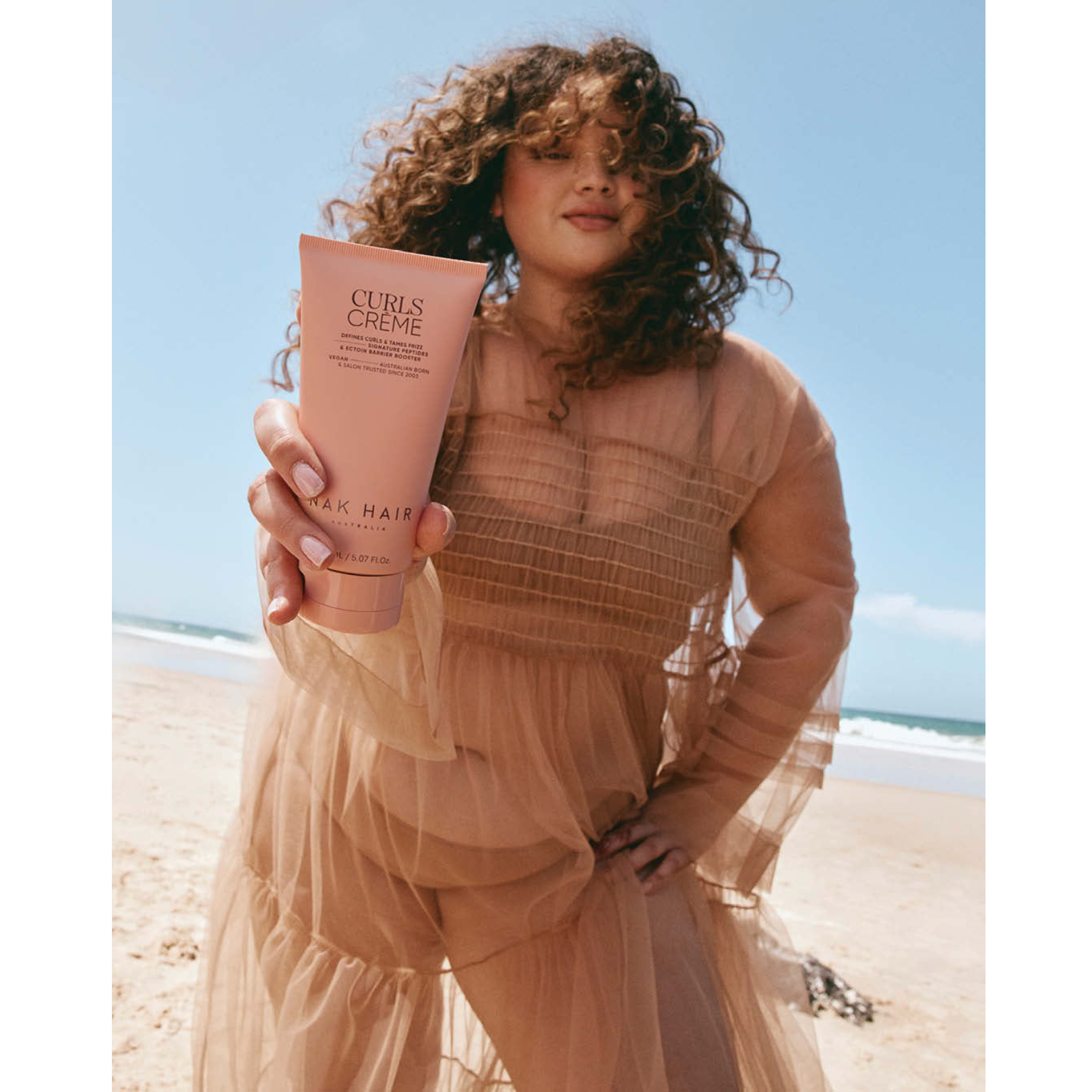 Woman with bouncy, defined curly hair holding NAK Hair Curls Crème 150ml vegan anti-frizz curl cream on a beach