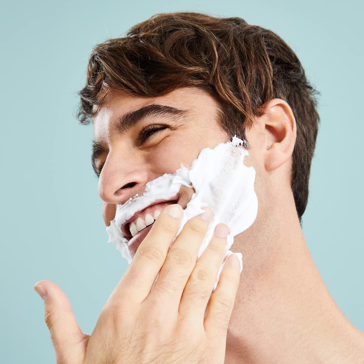 Smiling man applying Nivea Men Fresh Kick Shaving Foam 200ml to his face against a pale blue background, demonstrating its refreshing foam coverage before shaving.