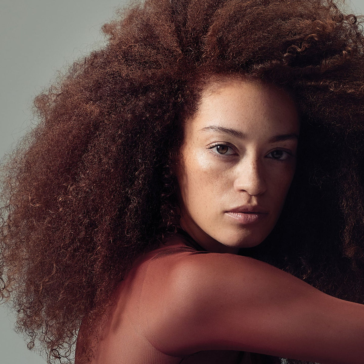 Model with voluminous, defined brown curls against a muted grey background, demonstrating lightweight texture and touchable hold.
