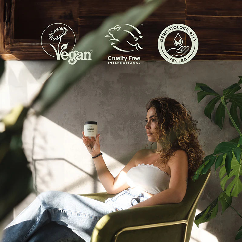 Woman holding a curl jar in a sunlit room with 'Vegan', 'Cruelty Free International', and 'Permanently Tested' logos.