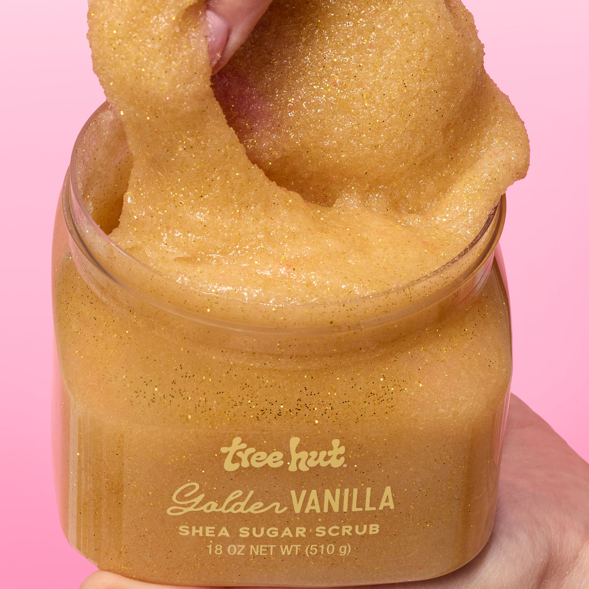 hand picking up Tree Hut Golden Vanilla Shea Sugar Scrub 18oz out of tub, showcasing consistency when applying product. on a pink background