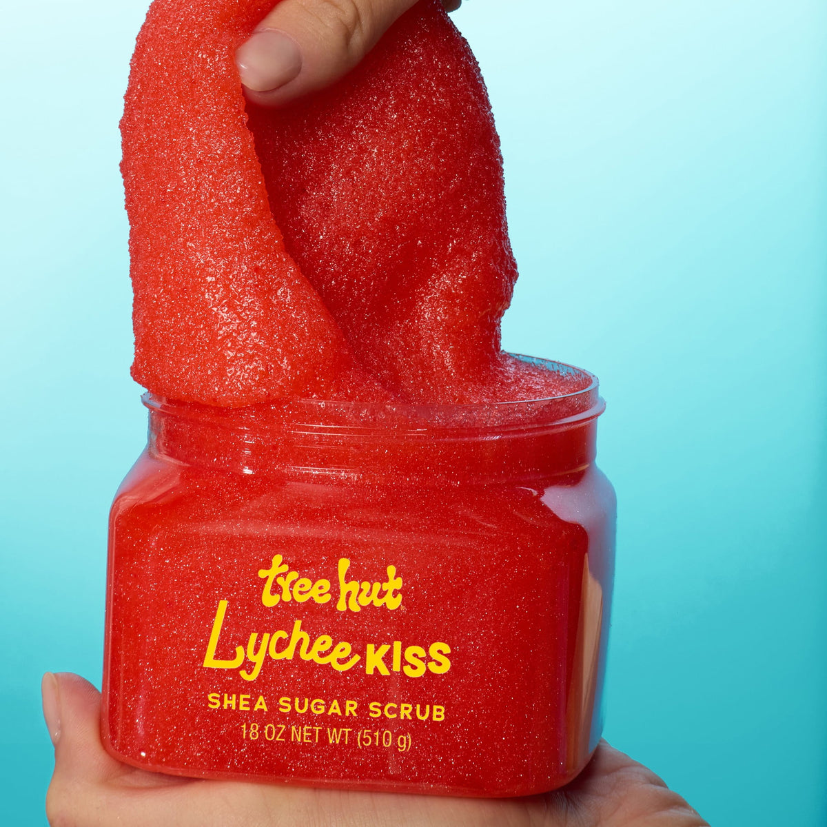 hand picking up Tree Hut Lychee Kiss Shea Sugar Scrub 18oz showing casing consistency when applying product on a light blue to solid cyan background