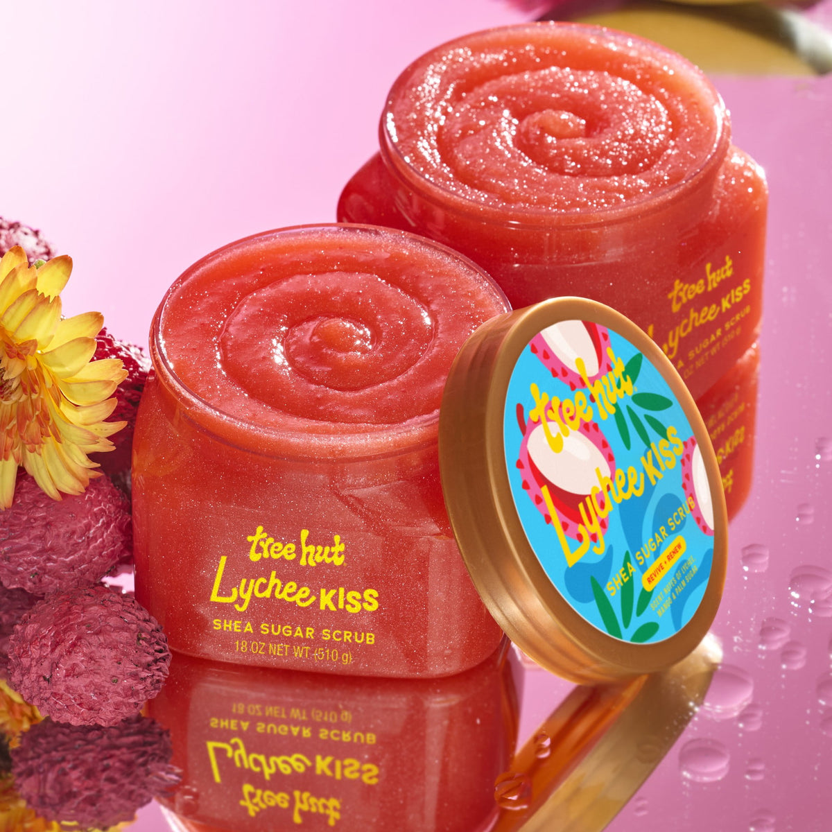 a picture of two tubs of Tree Hut Lychee Kiss Shea Sugar Scrub 18oz a lid leaning on the closest tub and the second without a lid with the lychee fruit on the left on reflective pink flooring