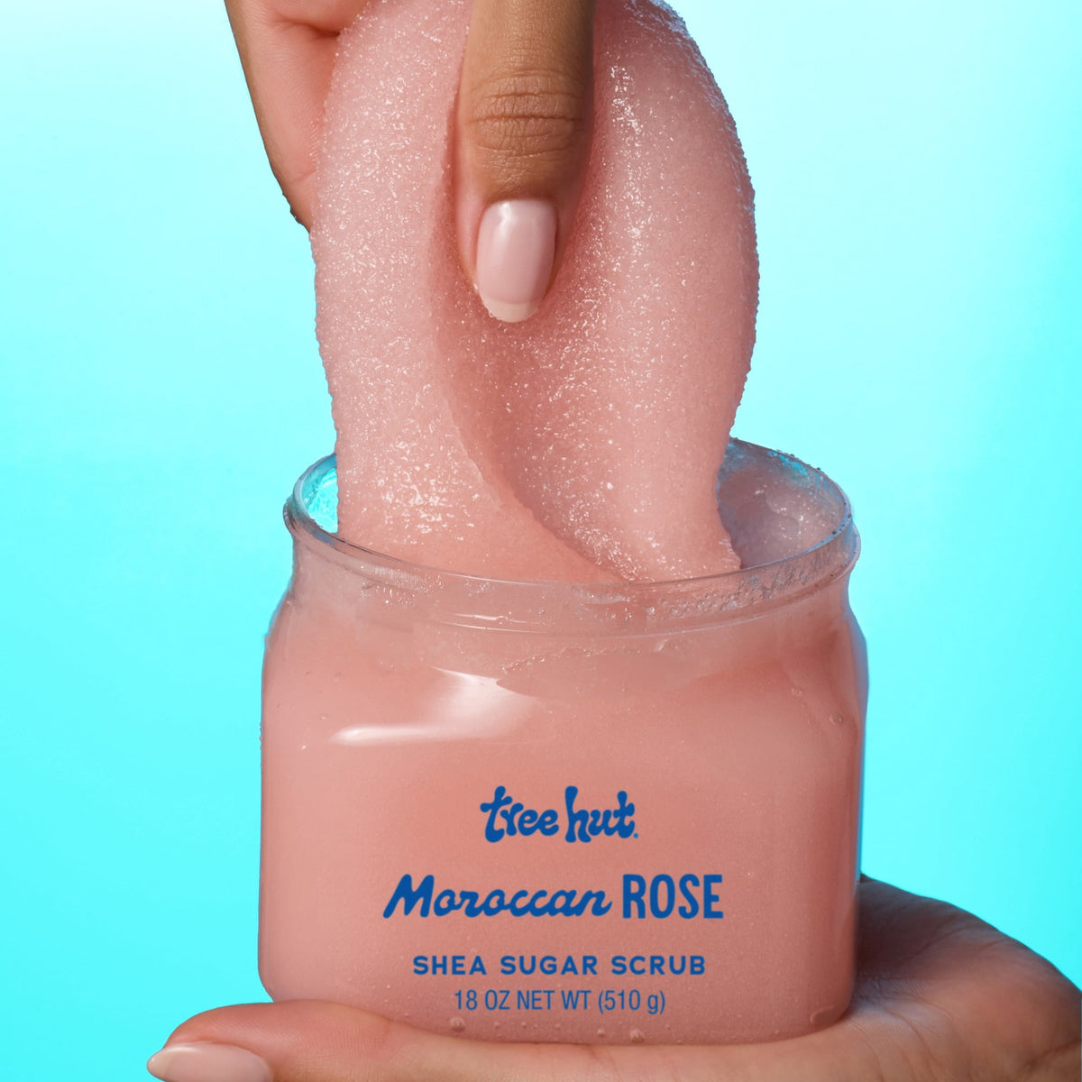 picture of hand picking up Tree Hut Moroccan Rose Shea Sugar Scrub 18oz, showcasing consistency when apply product on a light to solid blue background