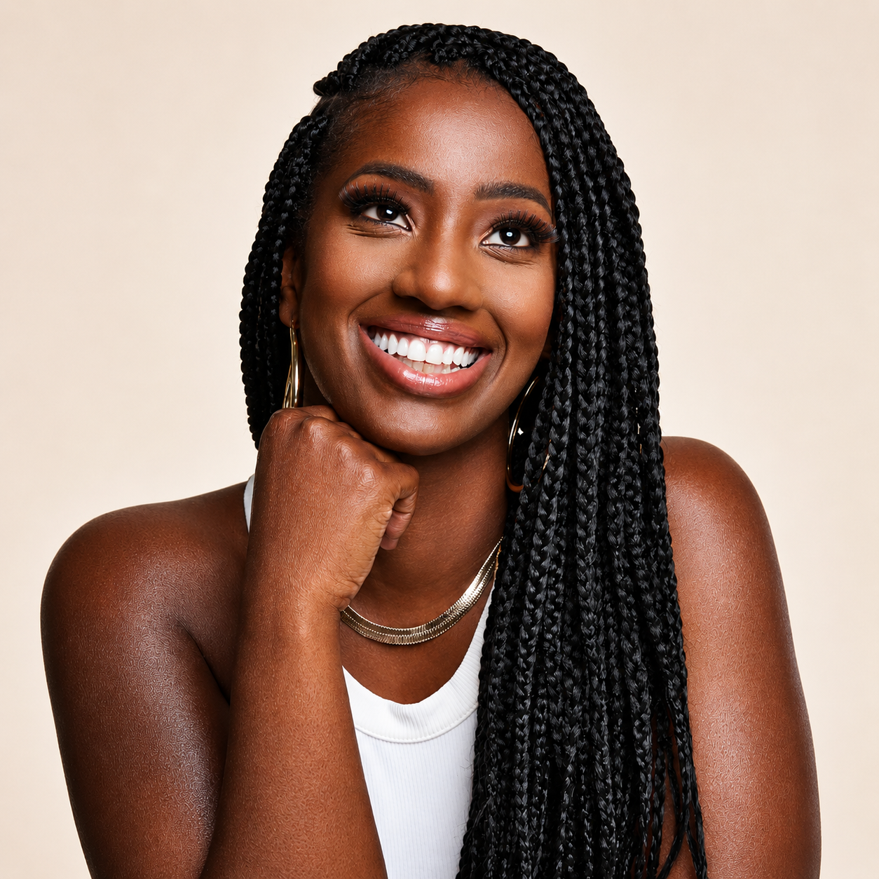 smiling woman with long black box braids wearing a white sleeveless top and gold hoop earrings, resting her chin on her hand against a soft neutral background.  portrait with natural skin texture, rich deep brown skin tone, bright smile, and studio lighting in square format.