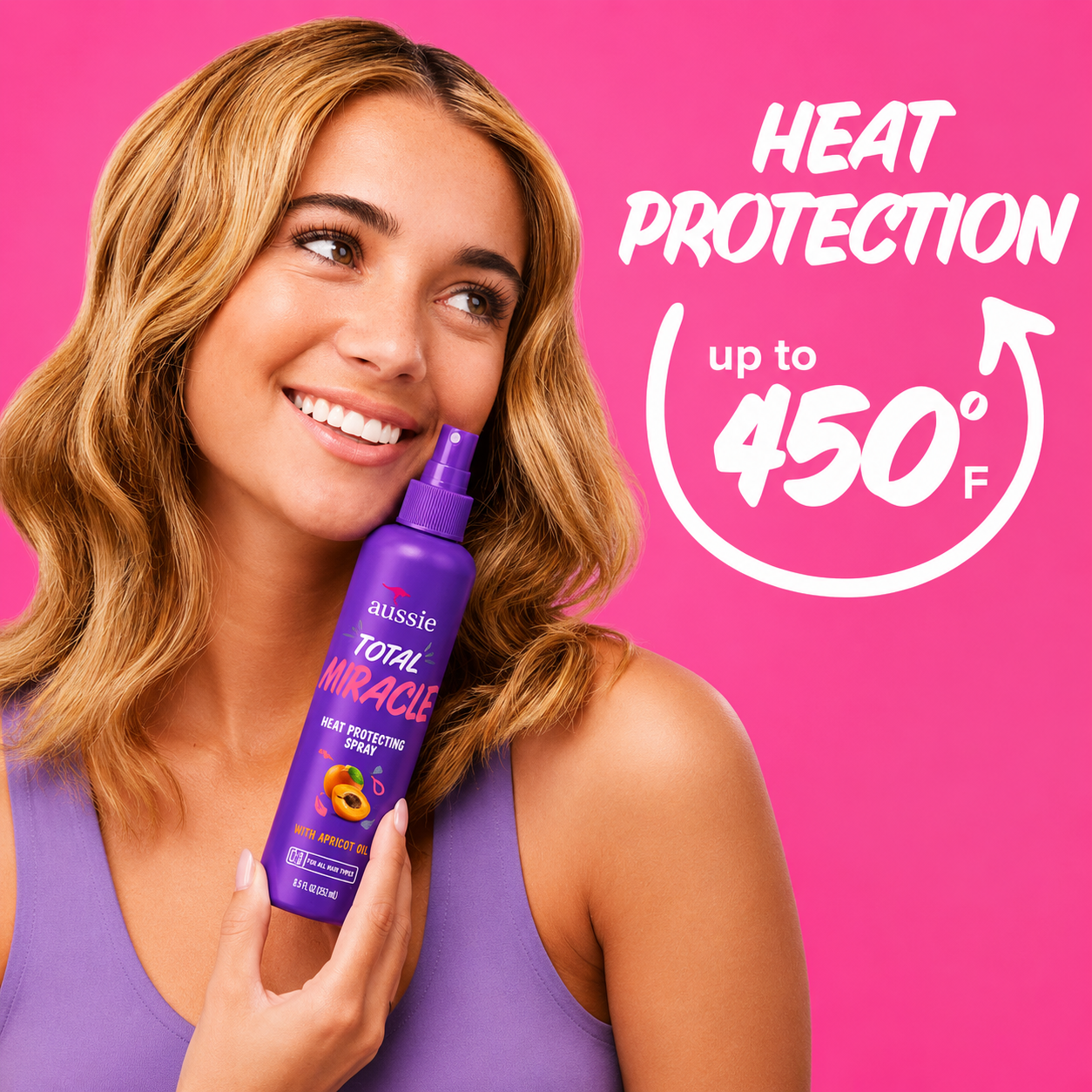 a promotional image featuring a smiling woman holding aussie total miracle heat protecting spray against a bright pink background. bold white text highlights heat protection up to 450°f, showcasing protection from heat styling while helping keep hair soft, smooth, and healthy-looking.