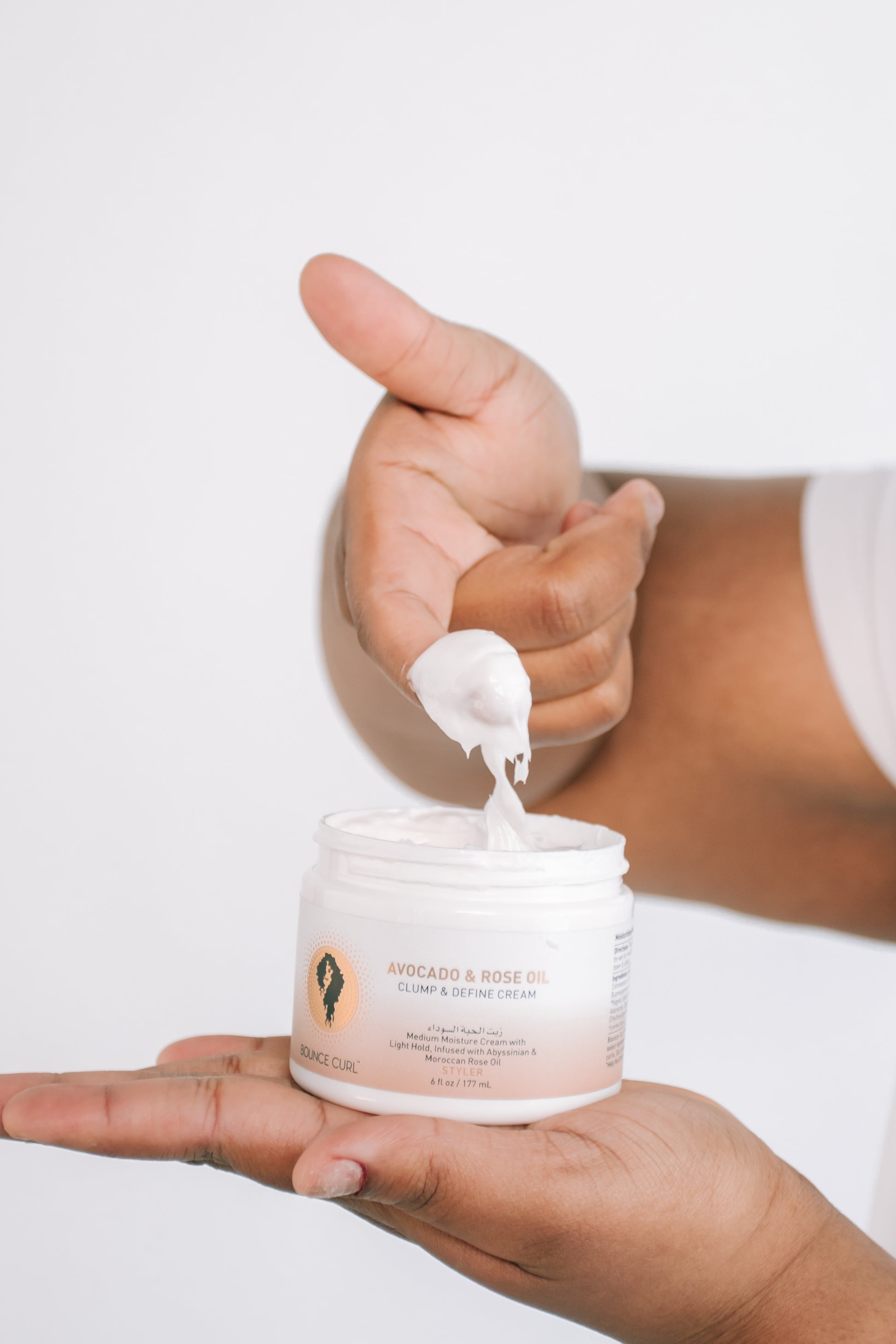 A person's hands holding an open white jar of bounce curl avocado & rose oil body and skin cream, with a dollop of the white cream being lifted by a finger against a clean white background.