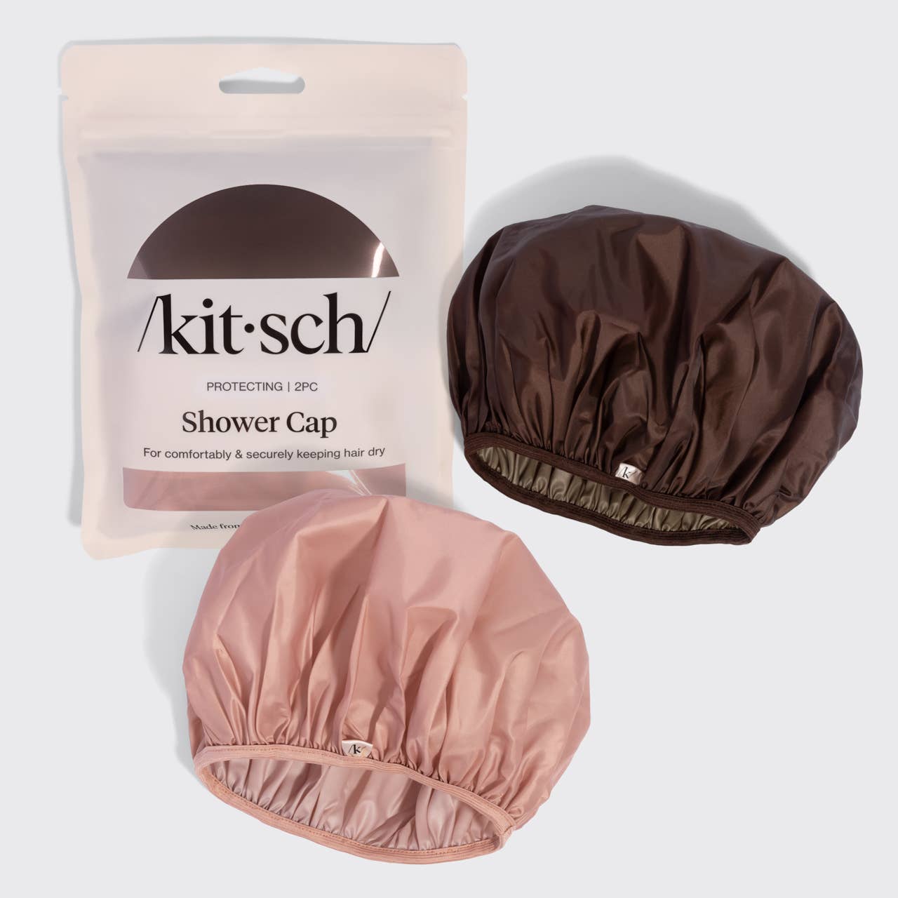 Two kitsch satin shower caps shown with their retail pouch packaging on a light gray background. One shower cap is a dusty pink color and the other is a dark brown color.