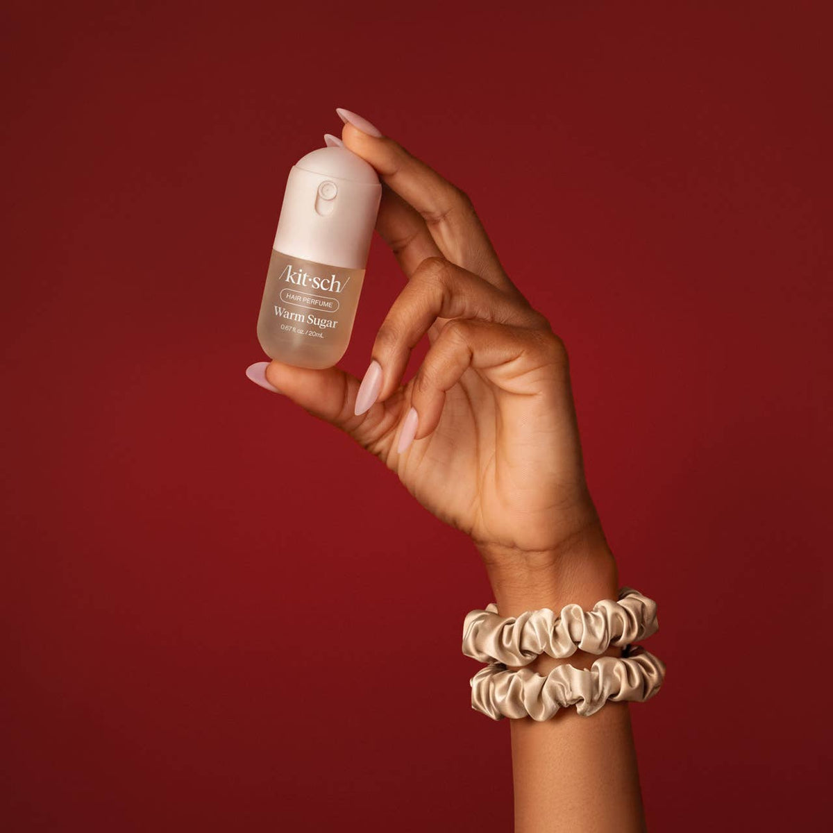 A hand holding a small bottle of Kitesch Haircare Minoxidil against a solid dark red background, with two cream satin scrunchies on the wrist.