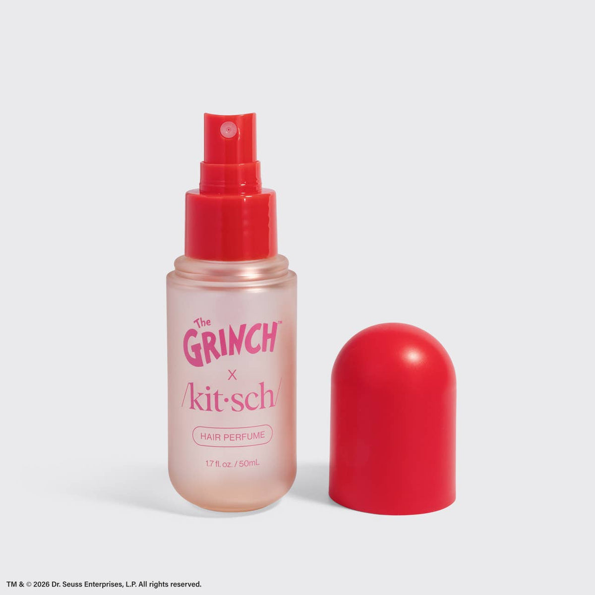 A bottle of Dr. Seuss Enterprises x Kitsch Hair Perfume with a translucent pink body and a bright red spray top, shown next to its matching red cap on a light gray background.
