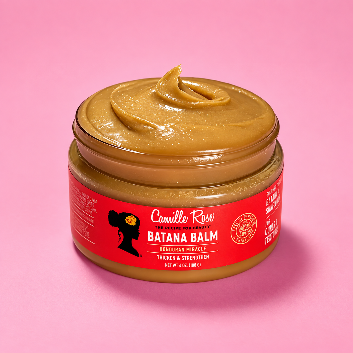 A jar of Camille Rose Batana Balm with a thick brown paste visible at the open top, set against a solid pink background.