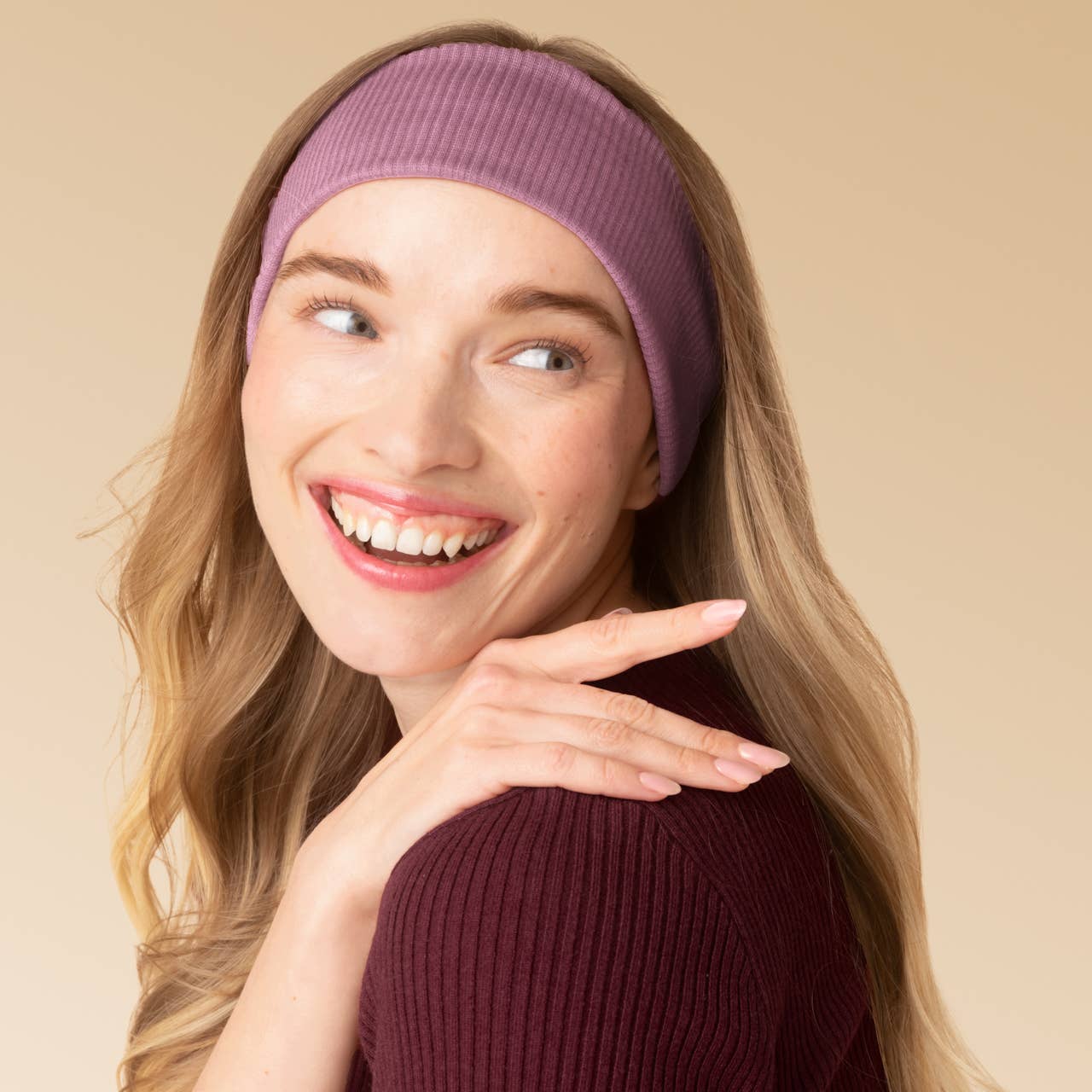 A smiling woman with long blonde hair wears a wide ribbed mauve-purple headband and a dark burgundy ribbed top against a plain beige studio background.
