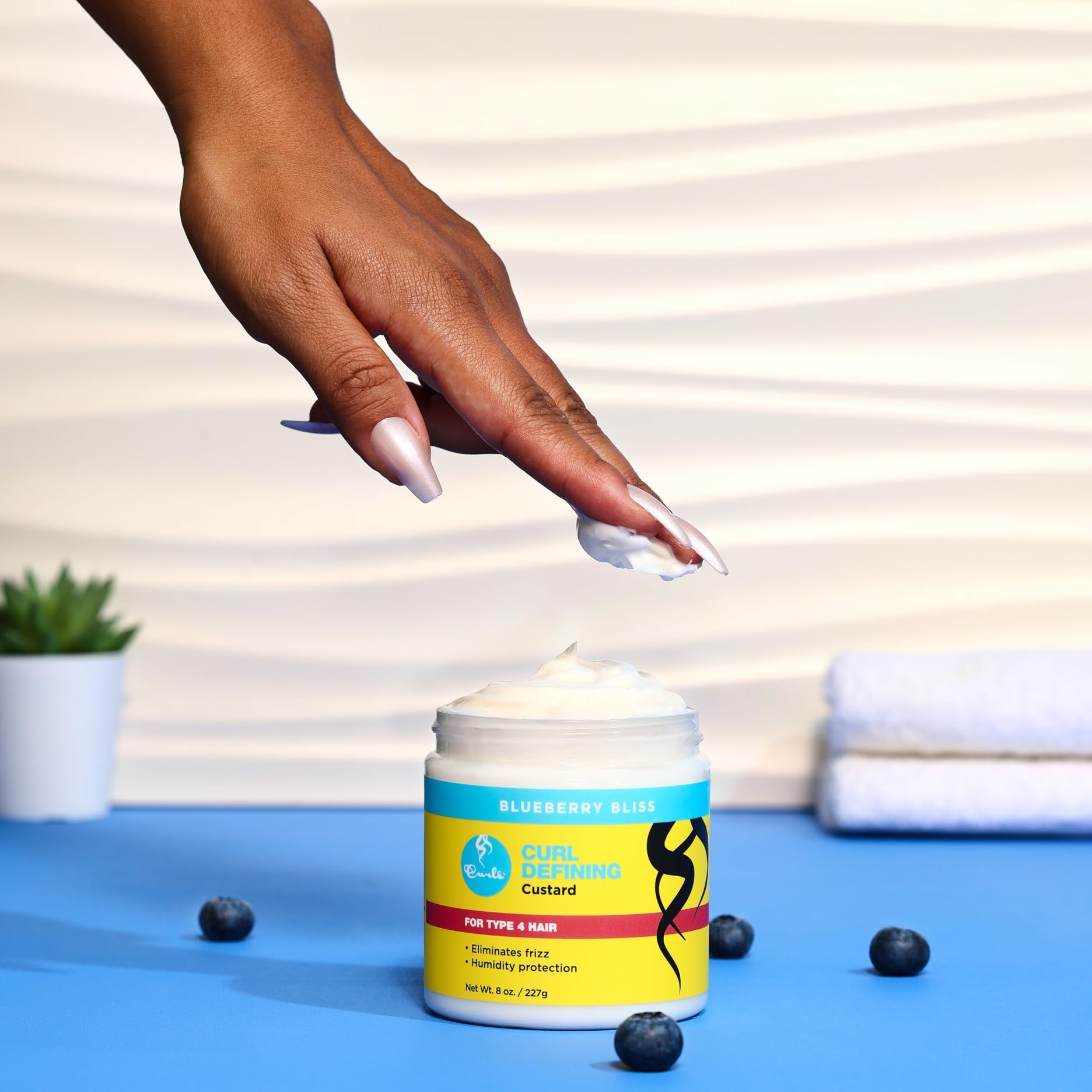 Hand reaching for a container of Curls Blueberry Bliss Defining Custard - 8 oz, Cream for Frizz Control & Humidity Protection, Enriched with Aloe Leaf Extract, Coconut Oil, Shea Butter & Mango Seed Butter, Type 4 Coils
