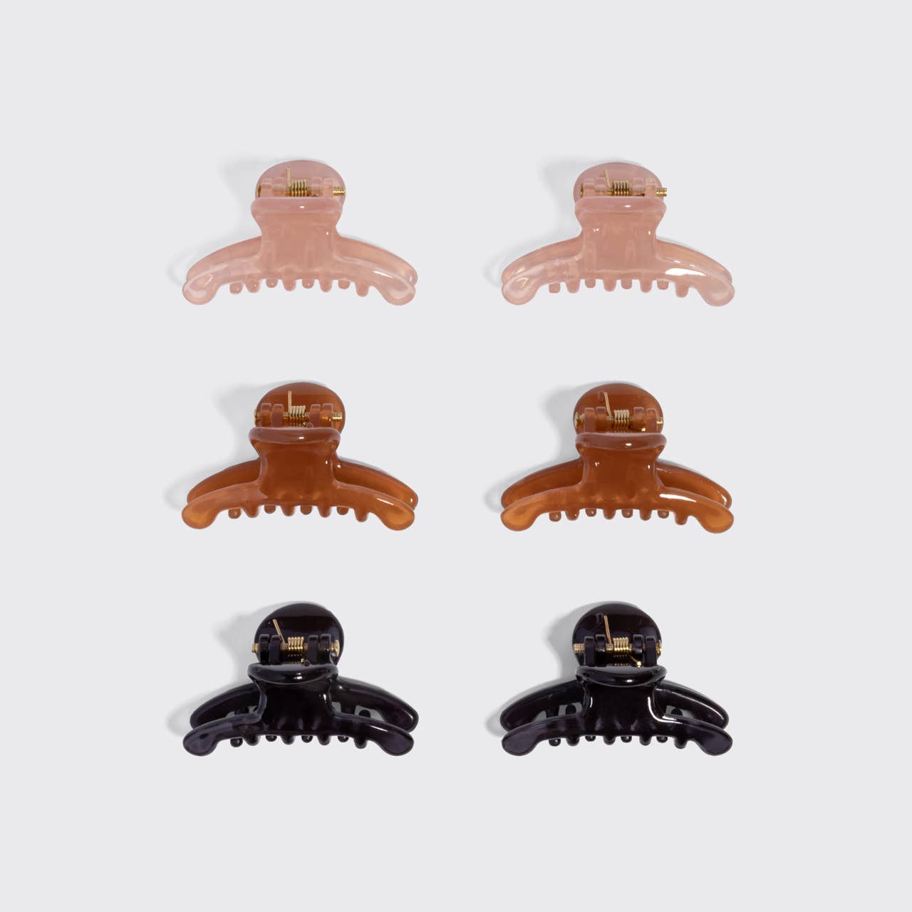 Six hair claw clips arranged in two columns of three on a light gray background. The clips come in translucent pale pink, warm amber-brown, and glossy black finishes, each with a small gold metal accent on the inner band.